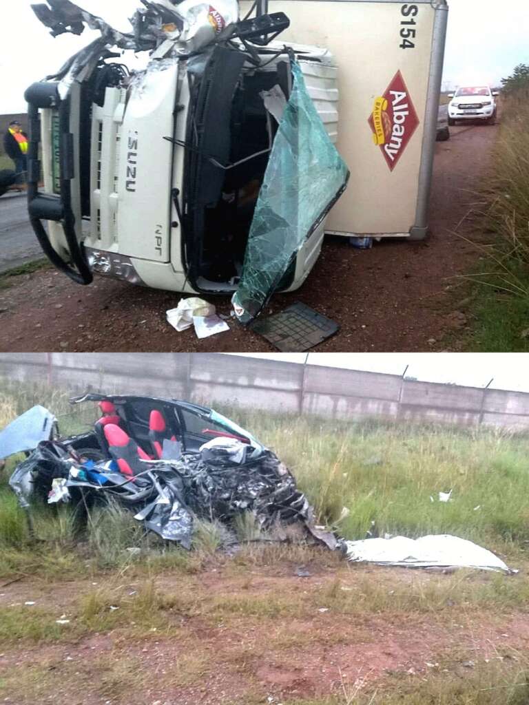 Overturned delivery truck with a shattered windshield on a roadside. Nearby, a severely damaged car with red seats sits in grassy terrain, depicting an accident scene.