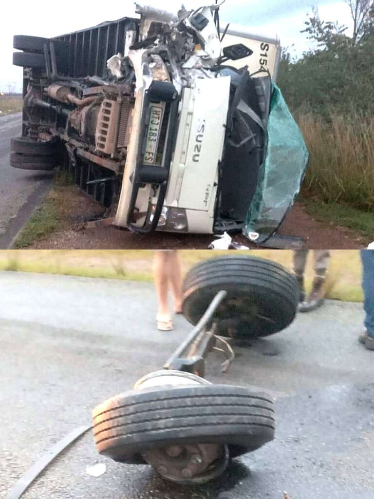Overturned white truck lies on its side on a rural road, with visible debris and a damaged front. Nearby, detached wheel axle rests on wet pavement.