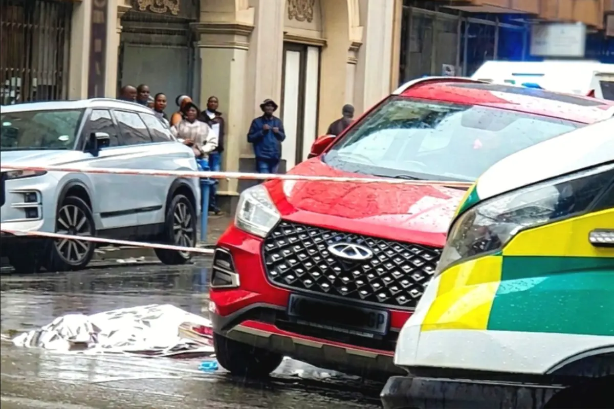 A rainy urban scene showing a red SUV and a white and green emergency vehicle on a wet street. A foil-covered object lies on the ground, with police tape and onlookers in the background.