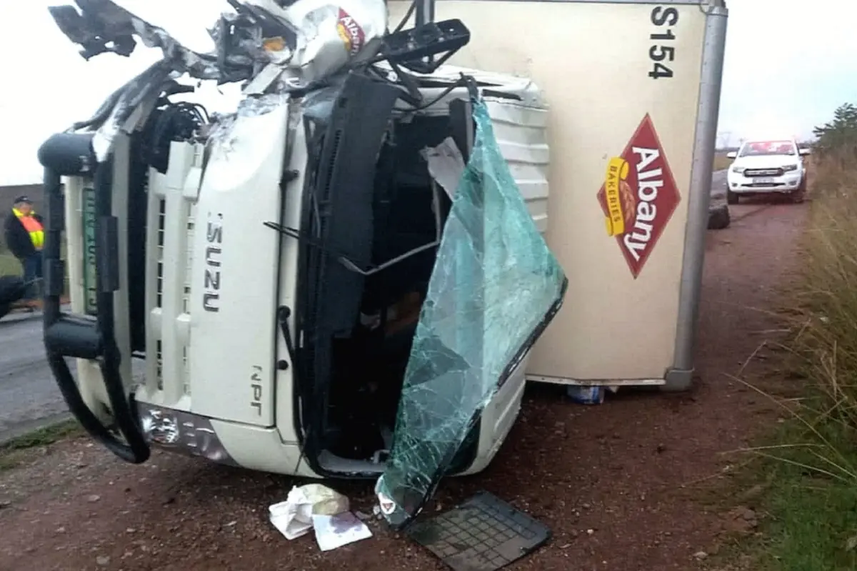 Overturned Isuzu truck on its side, damaged front and broken windshield. The truck carries Albany branding. Emergency vehicles in the background.