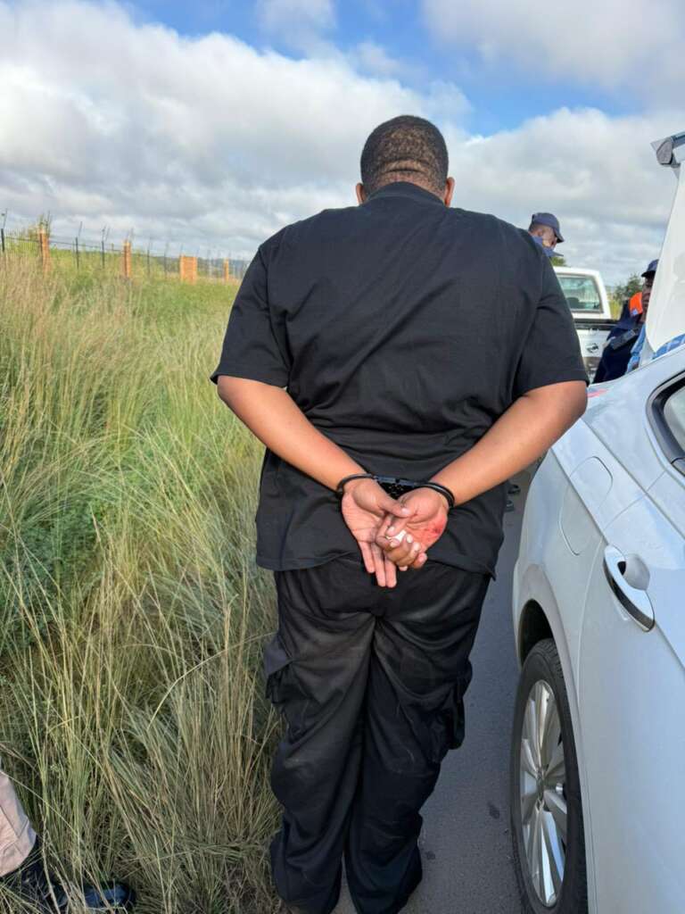A person in a black outfit stands handcuffed on a road beside a white car, facing away, next to grassy roadside under a cloudy sky.