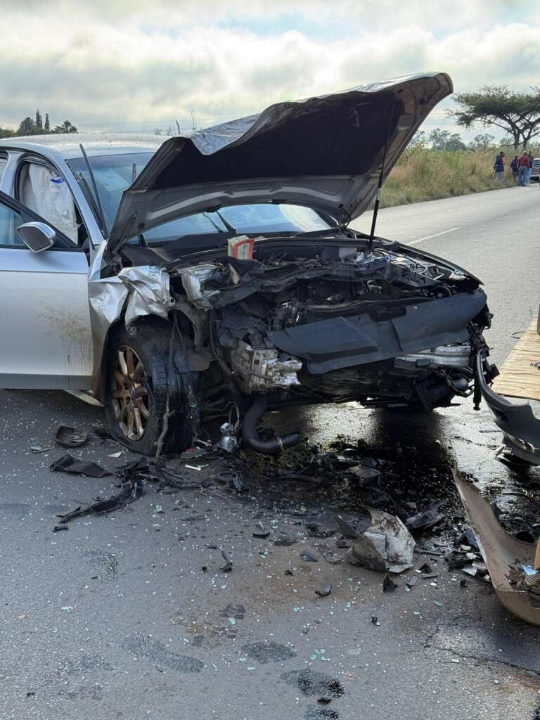 A damaged white car with a crumpled front and raised hood sits on a road lined with debris and shattered glass, suggesting a recent accident.