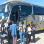 People wait in line to board a gray bus labeled "Delta" on a clear day. The scene conveys anticipation and travel.
